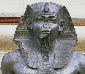 Busto esculpido em pedra escura de um faraó, sendo que o nariz está quebrado.