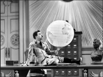 O Grande Ditador Chaplin com uniforme militar segurando um globo terrestre e sentado numa mesa.