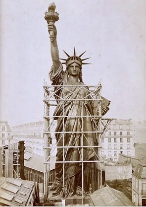 Foto antiga de uma estátua grande de mulher com uma coroa na cabeça e segurando um tocha na mão direita com o braço erguido.