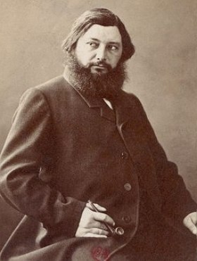 Foto em sépia de um homem de mais ou menos 40 anos de cabelo liso e barba, sentado e usando uma roupa escura
