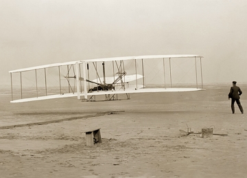 Primeiro voo dos irmãos Wright em 1903 Foto em preto e branco mostrando um pequeno avião levantando voo e um homem ao lado